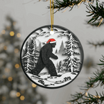 Round Christmas ornament featuring a black Bigfoot silhouette wearing a Santa hat, walking through a snowy forest scene.