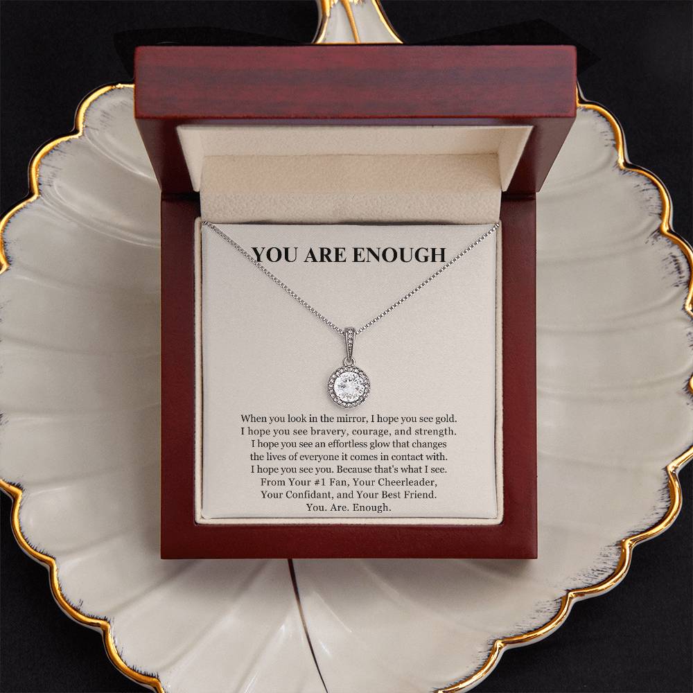 The Eternal Hope Necklace - You Are Enough, featuring a silver round pendant, is displayed in an open wooden box with YOU ARE ENOUGH on the lid and a self-love message inside. The box sits on a gold-trimmed decorative plate.