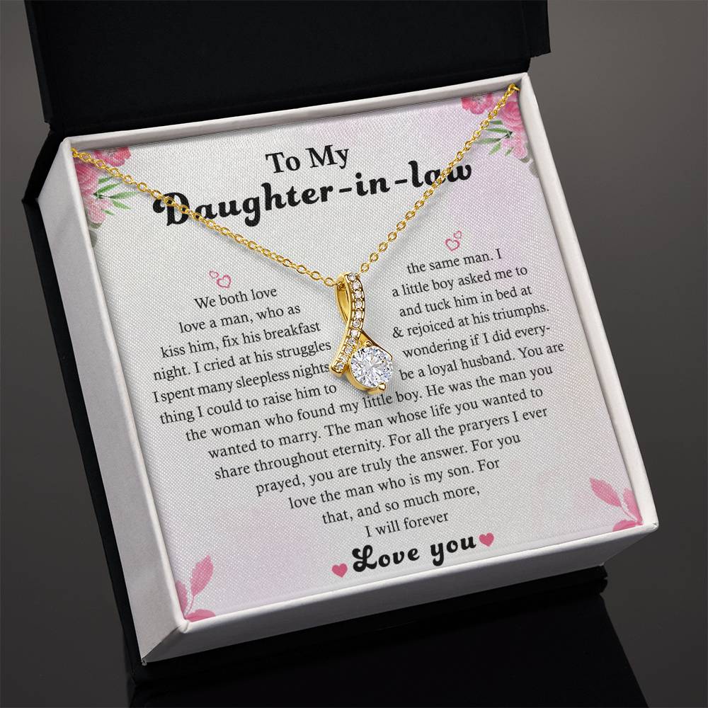 The To My Daughter-in-Law Alluring Beauty necklace, featuring a ribbon-shaped pendant in 14k white gold finish, is displayed in an open gift box adorned with pink flowers and includes a heartfelt message card.