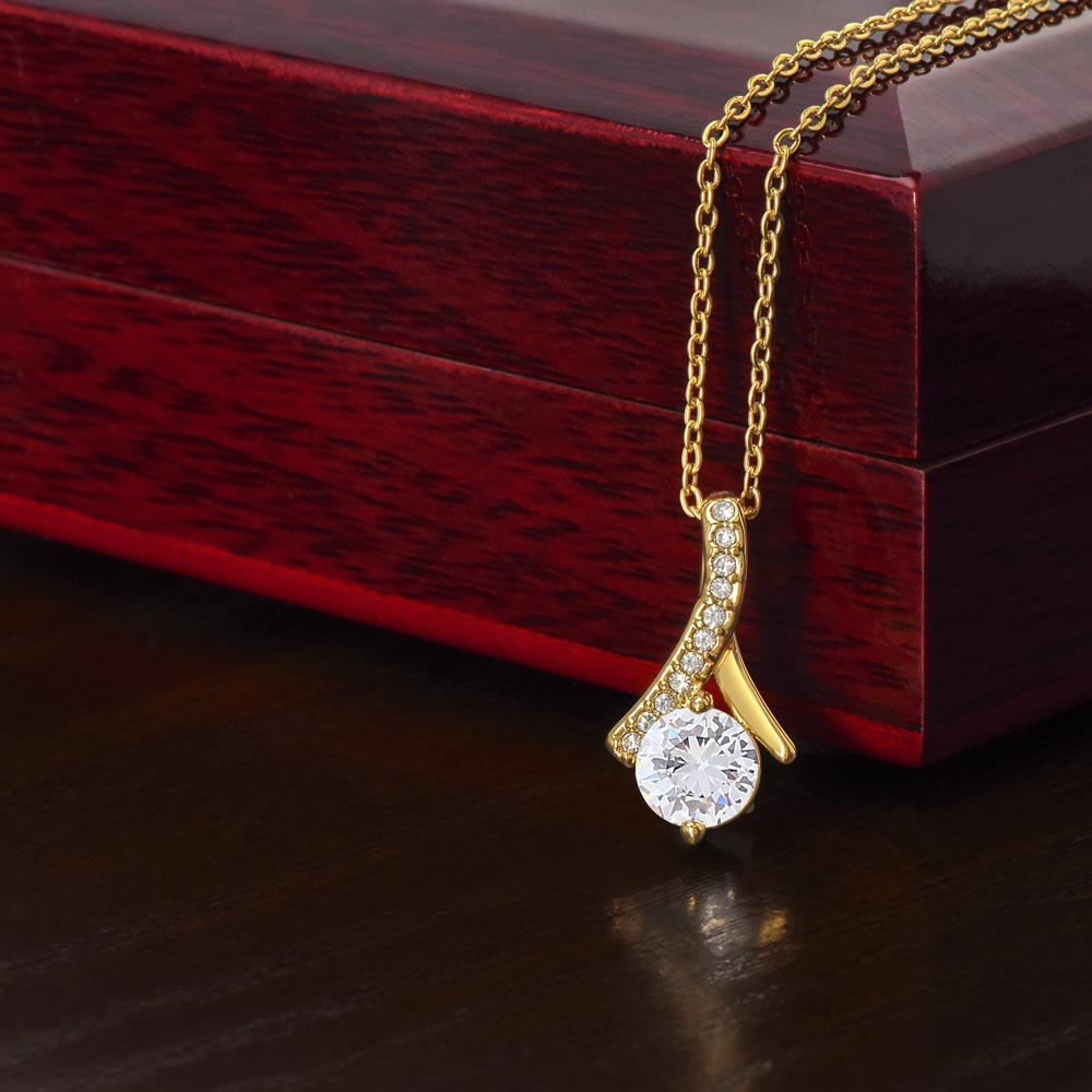 The To My Daughter-in-Law - Alluring Beauty necklace with message card rests on a polished wooden box, featuring a gold pendant with a large round clear gemstone and small embedded stones, elegantly showcasing its ribbon-shaped charm against the dark surface.