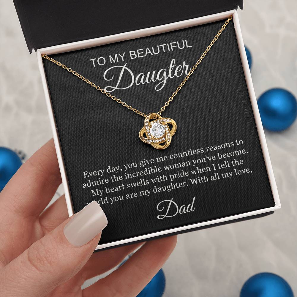 A hand holds an open box displaying the To My Beautiful Daughter | Love Knot Necklace in 14k white gold with cubic zirconia. The box features a heartfelt message from Dad, surrounded by blue ornaments.