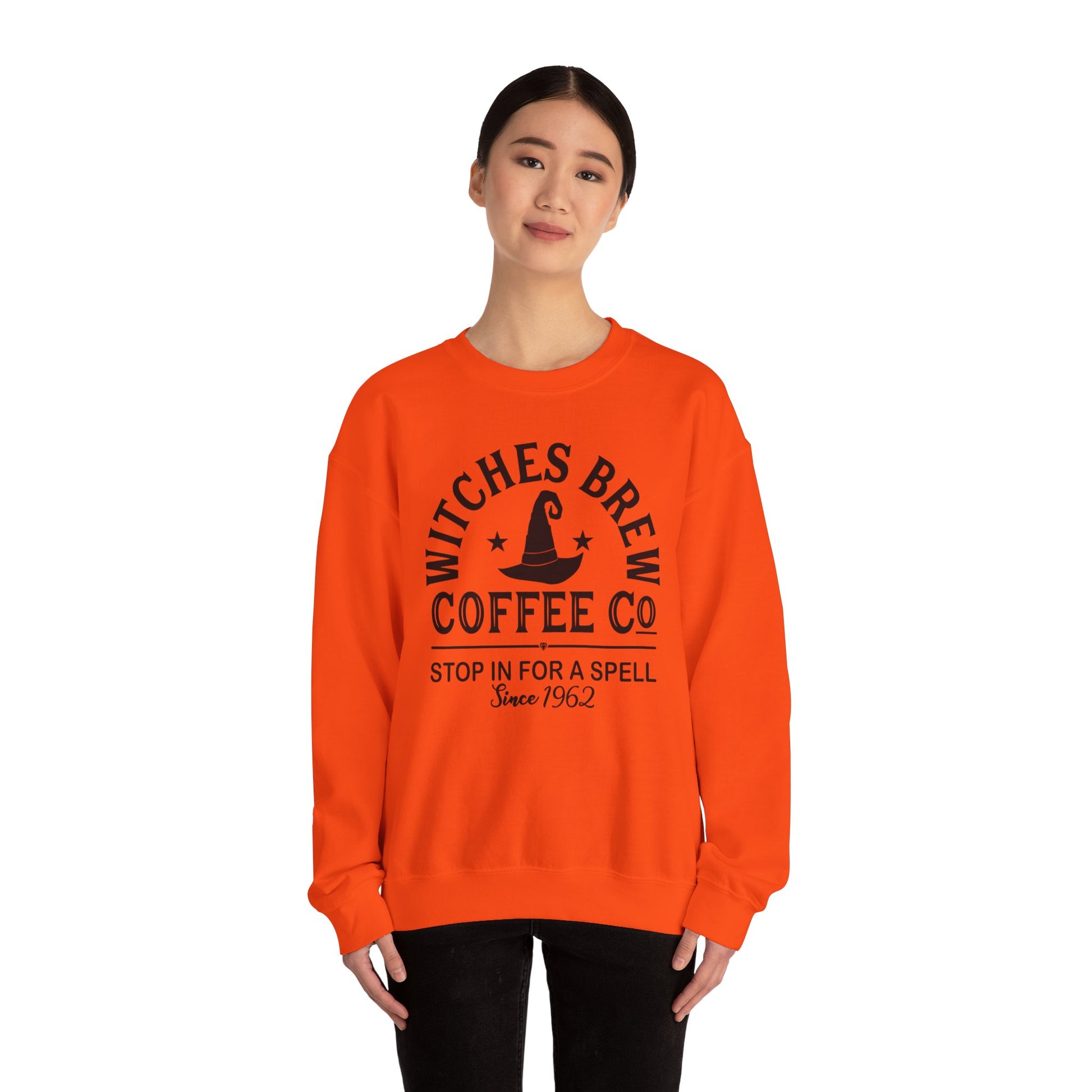 Wearing the Witches Brew Coffee Co. Sweatshirt - Stop in for a Spell! in vibrant orange, adorned with a witch hat design and dark hair pulled back, a person stands against a plain white background, showcasing the ultimate Halloween style.