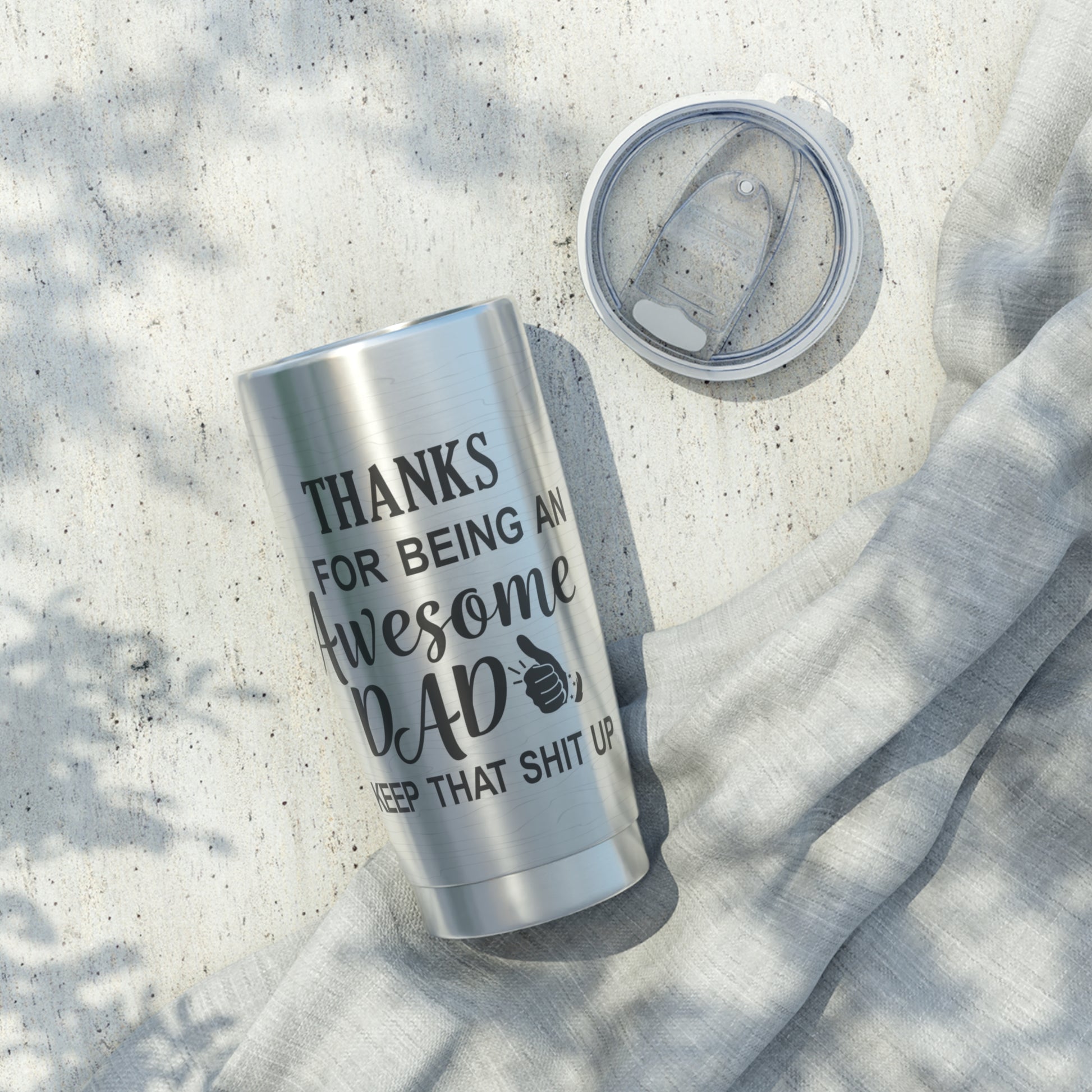 The “Thanks for Being an Awesome Dad” 20oz Stainless Steel Tumbler sits beside its clear lid on a light fabric, capturing sunlight—an ideal and unique Fathers Day gift.