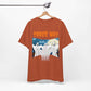 A rust-colored unisex jersey short-sleeve tee hangs on a hanger, showcasing three cartoon ghosts with THREE WAY in orange above them. The Three-Way Mirror Ghost Tee – A Haunting Twist on the Viral TikTok Trend, features trees, a moon, and is made from Airlume combed and ring-spun cotton.
