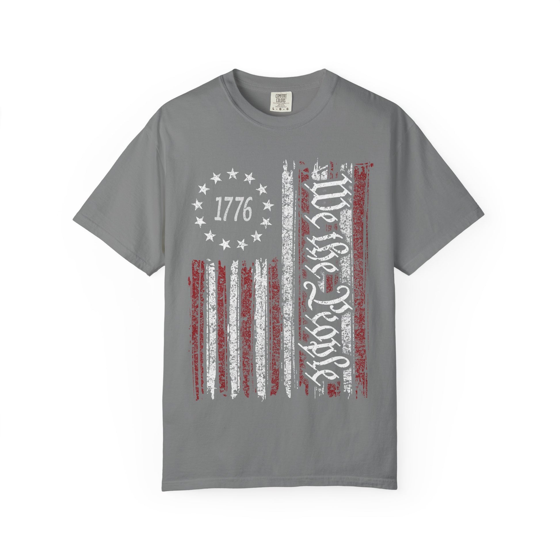 The We The People Distressed Flag Tee is a gray Comfort Colors 1717 unisex shirt featuring a distressed American flag with white script text and “1776” in a circle of stars on the upper left corner.