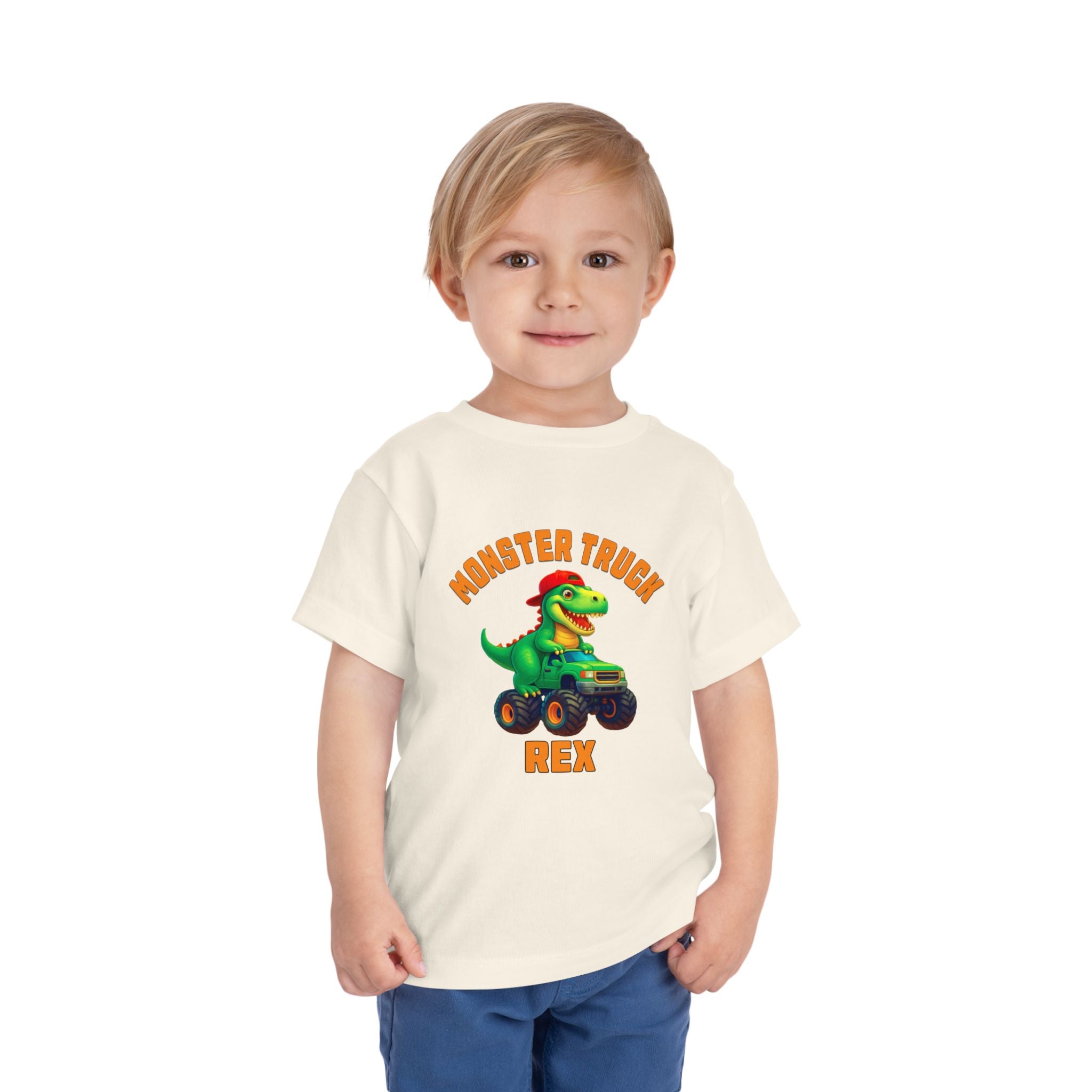 A young blonde child smiles in the Monster Truck Rex T-Shirt 🛻🦕 – Toddler Boys Tee, a white shirt with a cartoon dinosaur driving a monster truck and Monster Truck Rex text, standing against a white background.