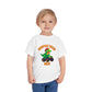 A smiling blonde-haired child wears the Monster Truck Rex T-Shirt 🛻🦕 – Toddler Boys Tee, featuring a cartoon dinosaur in a monster truck, and stands against a white background.