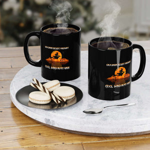 Two steaming black mugs with On a Dark Desert Highway designs exude spooky charm on a round tray with a spoon. Nearby, two macarons sit on a small plate, perfect for coffee lovers. The background is blurred for effect.