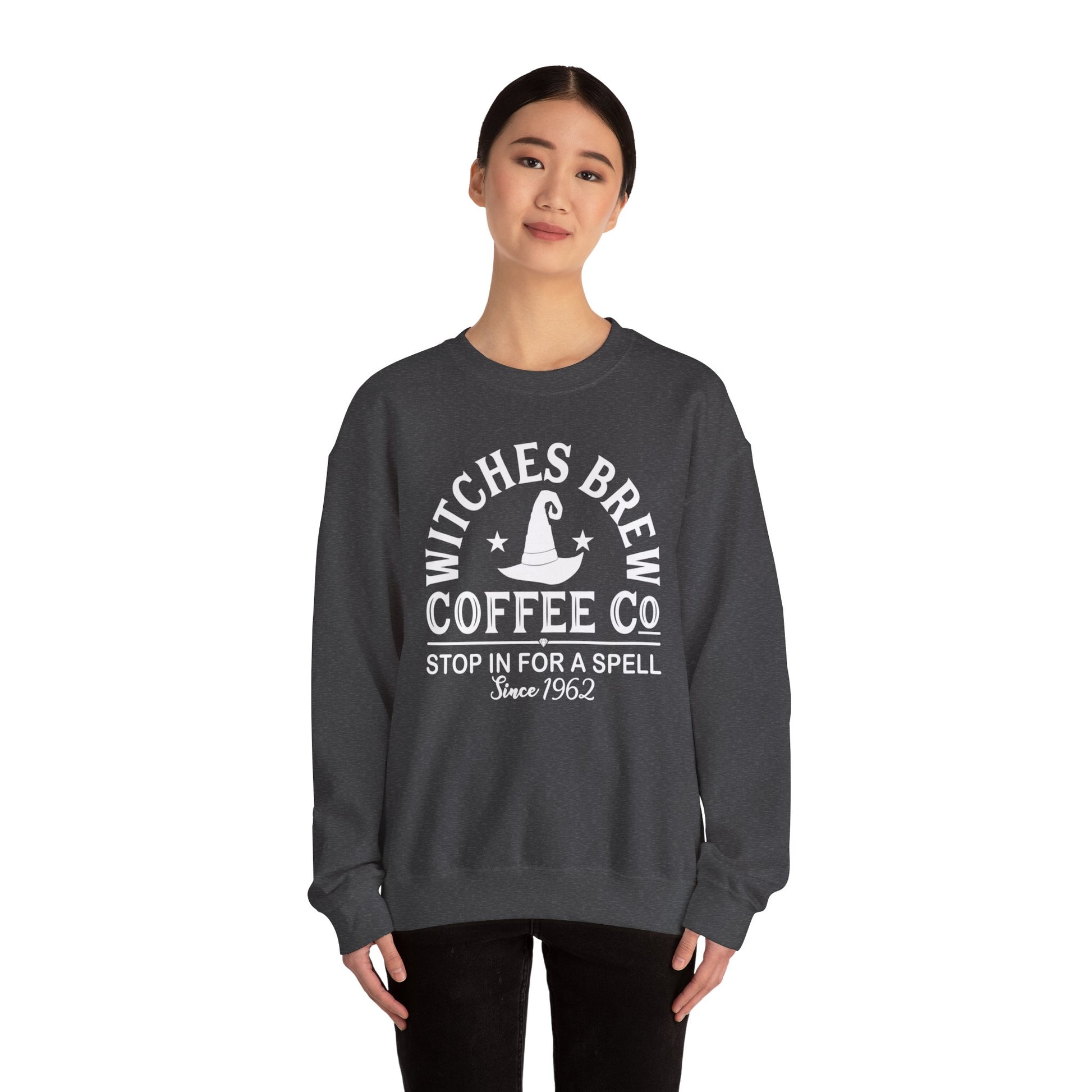 Wearing the Witches Brew Coffee Co. Sweatshirt - Stop in for a Spell! | Unisex Heavy Blend™ Crewneck Sweatshirt, featuring “Stop in for a spell—Since 1962” and a witch’s hat graphic, someone stands against a plain white background.