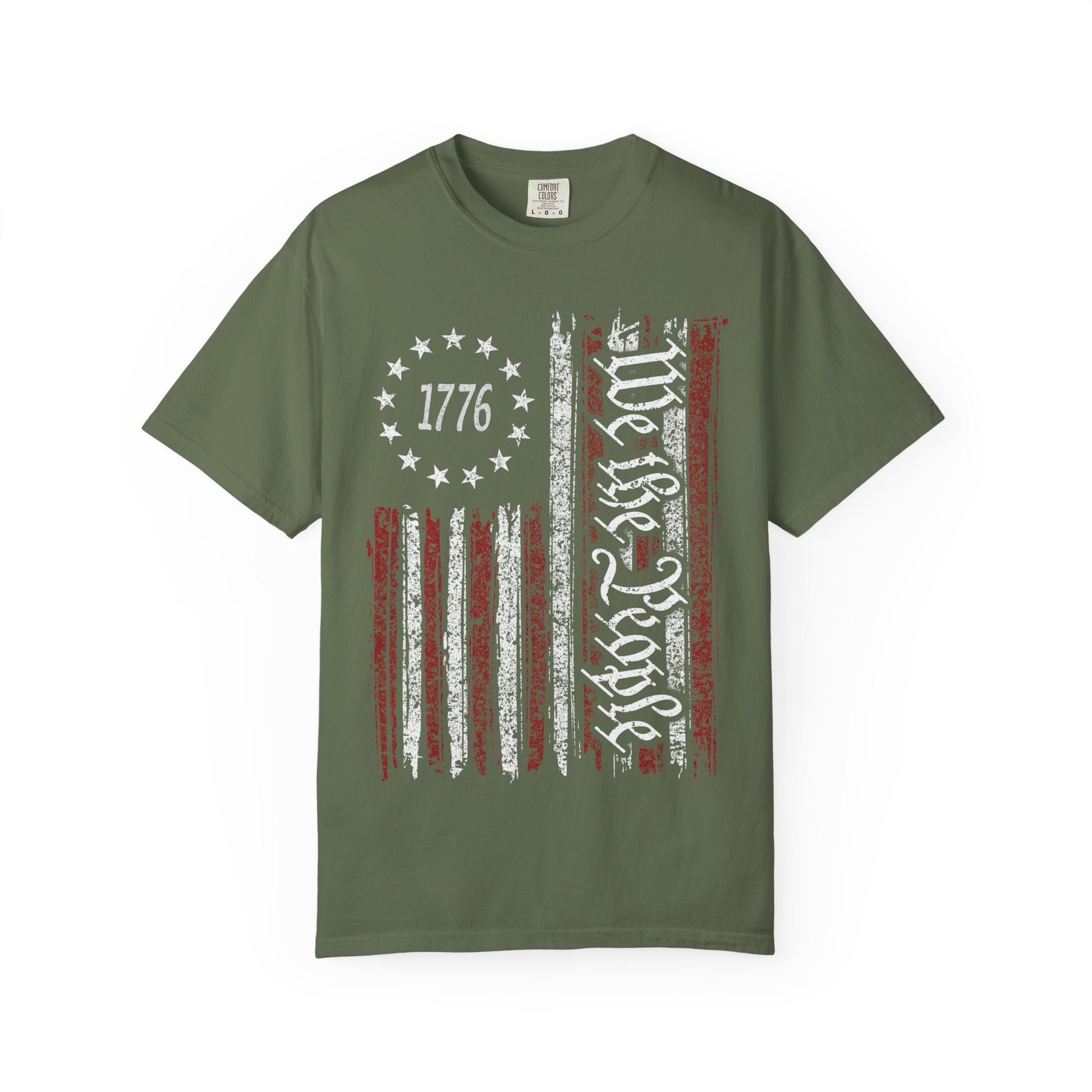 The We The People Distressed Flag Tee is an olive green Comfort Colors shirt with a weathered American flag, “1776” in the canton, and “Hold Fast” in white text across the stripes—a bold tribute to American freedom.