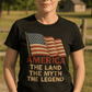 A woman with short blonde hair stands outdoors by trees and a wooden fence, wearing the America: The Land, The Myth, The Legend – 100% Cotton Tee featuring an American flag and patriotic text.