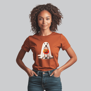 A woman, smiling spookily, wears a Ghostly Pup Halloween T-Shirt, featuring a playful ghost dog with a pumpkin bucket. She stands with her hands casually in her jeans pockets against a gray backdrop.