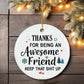 Round white Christmas ornament with a humorous appreciation message for an awesome friend, decorated with festive icons and hung on a holiday tree.