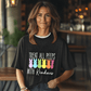Indoors, someone by a wooden counter wears the Treat All Peeps with Kindness! tee featuring colorful peep marshmallow bunnies and text. The softly blurred background highlights lights and furniture. This black unisex jersey is the perfect Easter tee for fun-loving souls!.
