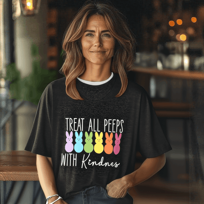 Indoors, someone by a wooden counter wears the Treat All Peeps with Kindness! tee featuring colorful peep marshmallow bunnies and text. The softly blurred background highlights lights and furniture. This black unisex jersey is the perfect Easter tee for fun-loving souls!.