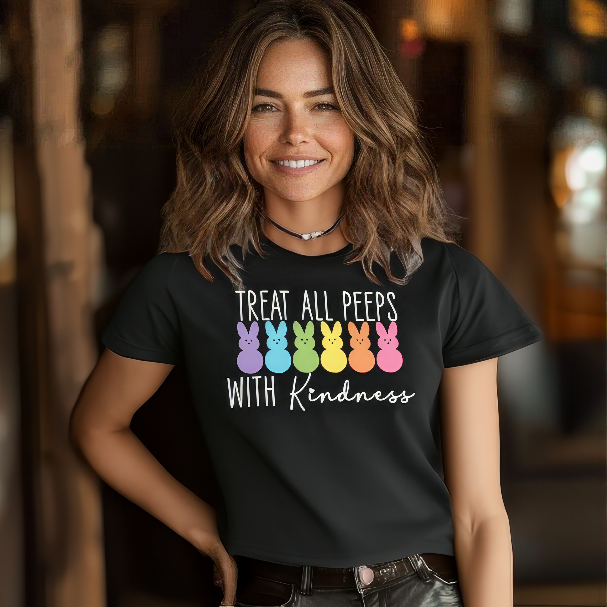 A woman smiles indoors, wearing a black unisex jersey tee crafted from soft Airlume combed cotton, adorned with colorful bunny silhouettes and the phrase Treat All Peeps With Kindness, known as the perfect Easter tee for fun-loving souls.