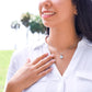 Smiling outdoors with blurred trees behind them, a person in a white shirt lightly touches their chest, where the Eternal Hope Pendant dangles in cubic zirconia set in 14k white gold, complemented by a heartfelt message card.
