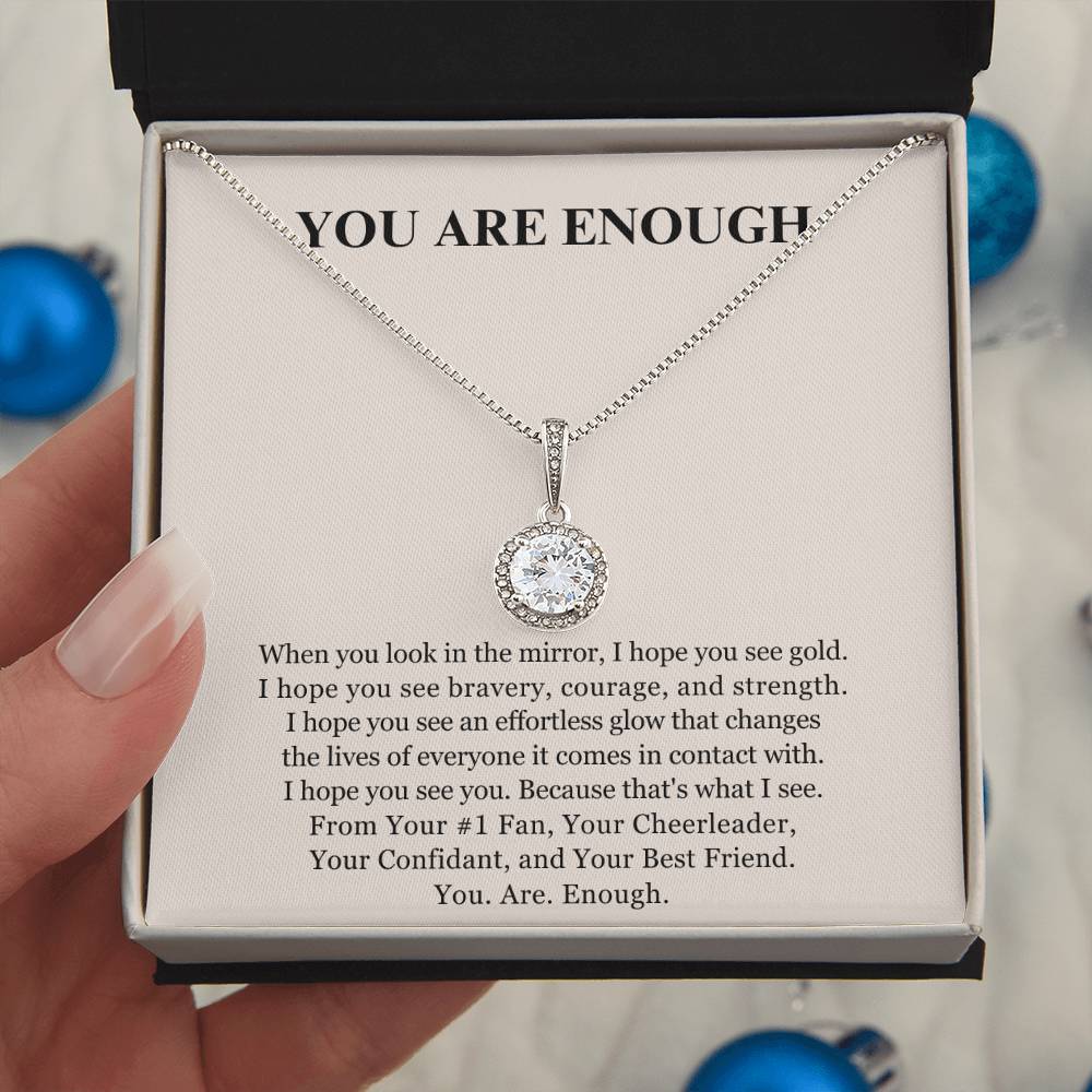 A hand holds an open jewelry box presenting the Eternal Hope Necklace - You Are Enough, a silver necklace with a round, sparkling pendant. Inside the lid, “YOU ARE ENOUGH” is inscribed above an uplifting message. Blue ornaments blur in the background.