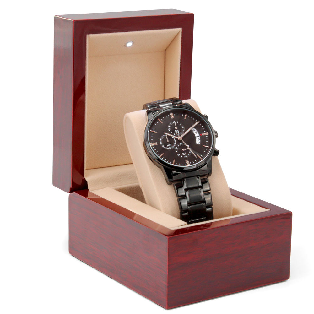 The Replacement Attachment, a sleek black metal wristwatch with a round face, is presented in an open wooden watch box with beige lining and a small LED light at the top.