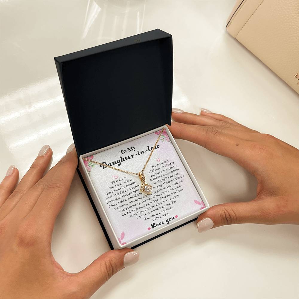 Hands presenting an open jewelry box containing the To My Daughter-in-Law - Alluring Beauty necklace with a cubic zirconia heart pendant. The accompanying message card begins with To my Daughter-in-Law and concludes with Love you, embellished with floral designs.