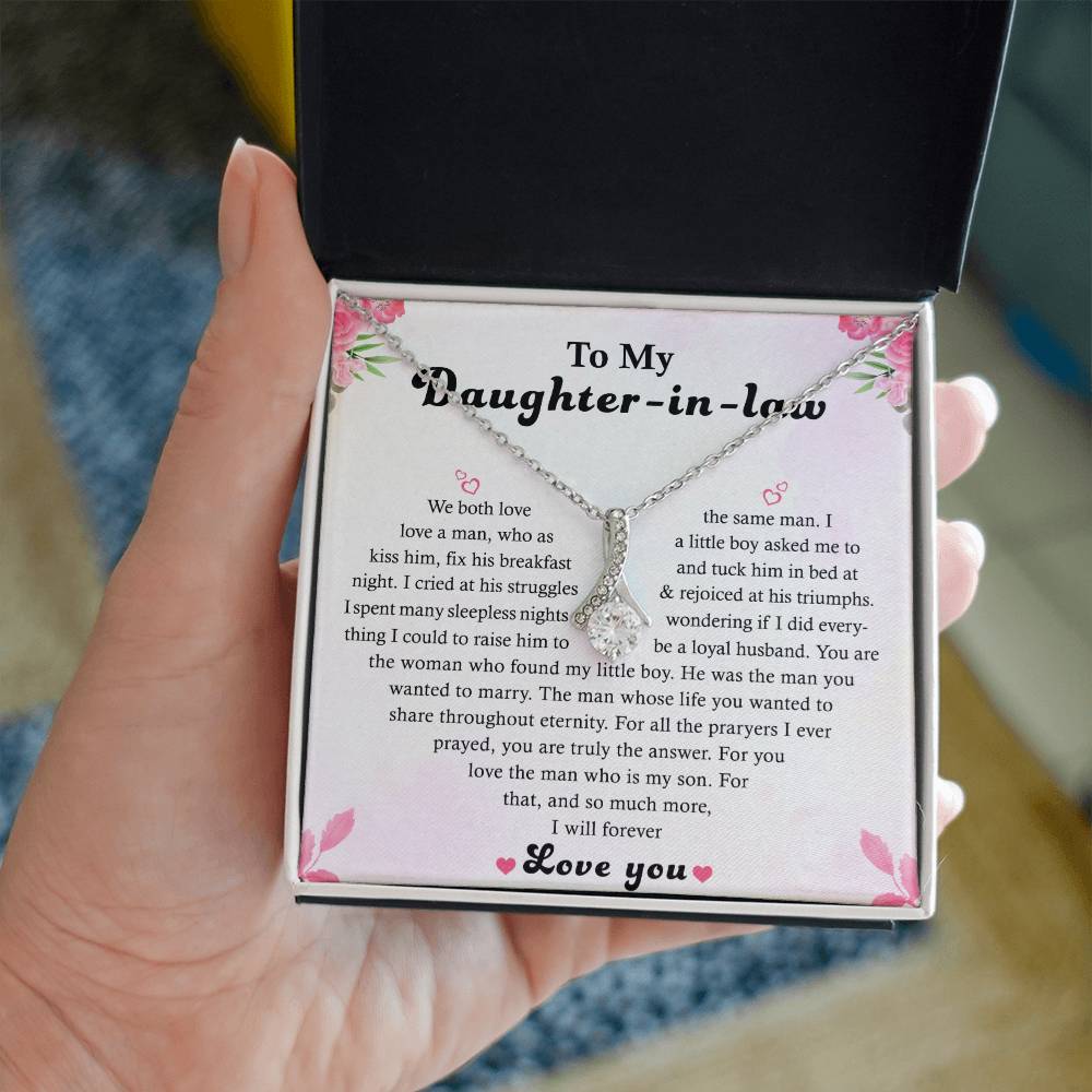 An open gift box displays the To My Daughter-in-Law - Alluring Beauty necklace featuring a ribbon-shaped pendant with a cubic zirconia center. The lid bears a heartfelt message to a daughter-in-law, adorned with floral accents, and highlights the necklaces elegant chain.