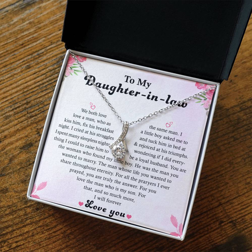 An open jewelry box on a wooden surface displays the To My Daughter-in-Law - Alluring Beauty necklace with a 14k white gold finish, featuring a tear-shaped pendant and a heartfelt message. Pink floral decorations surround the card.