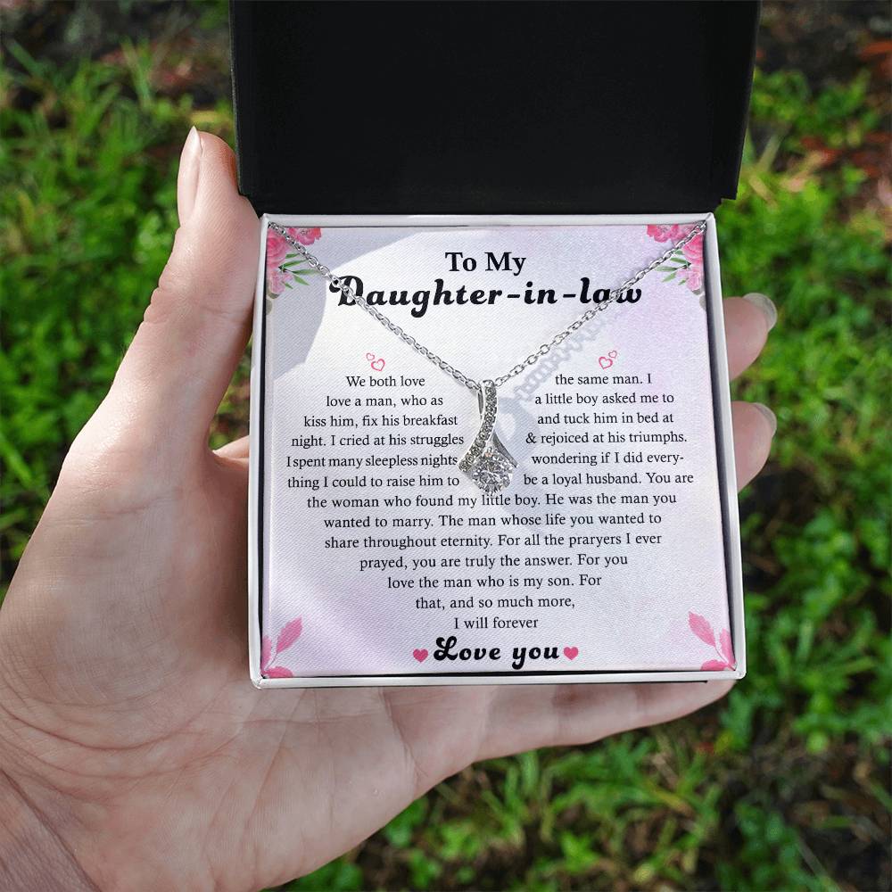 A hand presents an open box with the Alluring Beauty necklace, adorned with a floral design and 14k white gold finish. Inside the lid is a heartfelt message card titled To My Daughter-in-Law, expressing love and admiration. The box exudes elegance and sentiment.