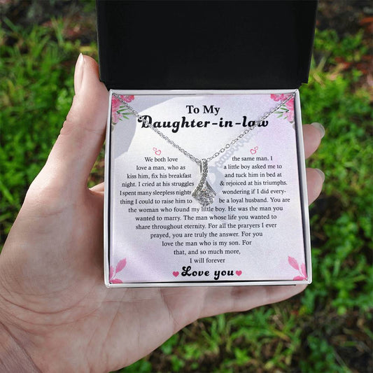 A hand presents an open box with the Alluring Beauty necklace, adorned with a floral design and 14k white gold finish. Inside the lid is a heartfelt message card titled To My Daughter-in-Law, expressing love and admiration. The box exudes elegance and sentiment.