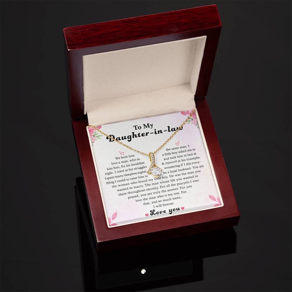 A wooden box with an open lid showcases the To My Daughter-in-Law - Alluring Beauty necklace, featuring a ribbon design adorned with sparkling cubic zirconia. Its presented on a card with the heartfelt message To My Daughter-in-law, decorated with pink flowers against a glossy black surface.