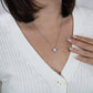 A woman in a white sweater displays the To My Daughter – Love Knot Necklace with Lion & Cub Message Card, featuring a square pendant near her collarbone. Her French-manicured hand gently touches her chest beside the necklace.