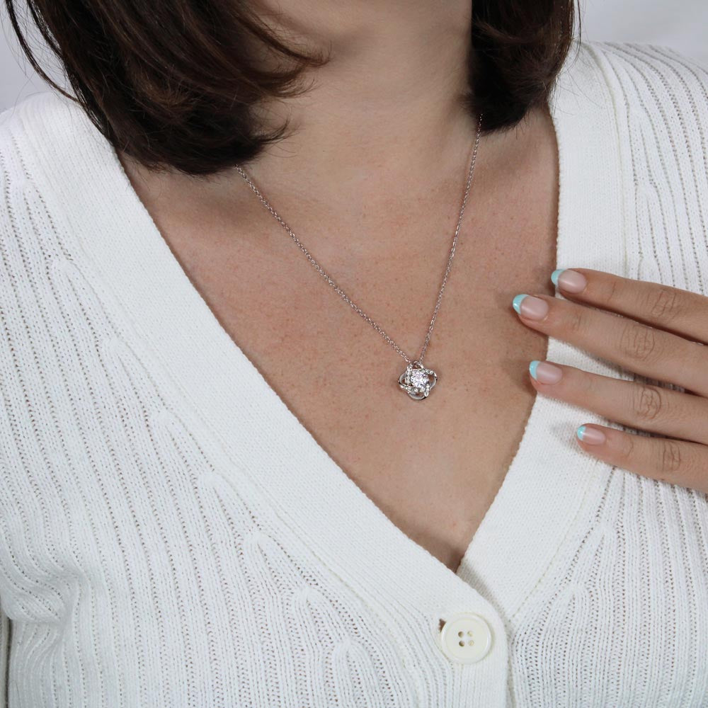 A woman in a white sweater displays the To My Daughter – Love Knot Necklace with Lion & Cub Message Card, featuring a square pendant near her collarbone. Her French-manicured hand gently touches her chest beside the necklace.