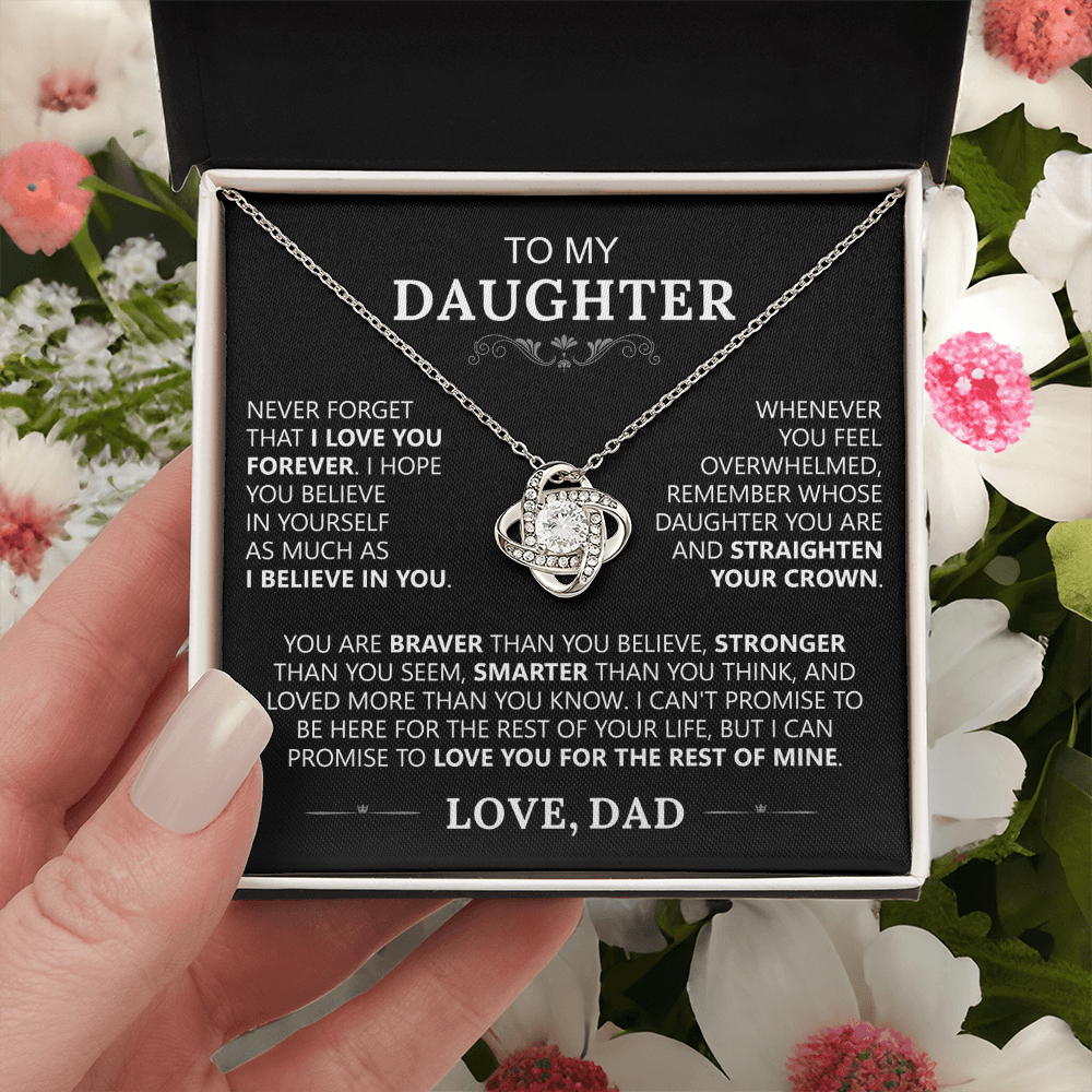 A hand holds an open box with the To My Daughter - Love Knot Necklace inside. The lid displays a heartfelt father-to-daughter message, symbolizing their lasting bond, against a background of pink and white flowers.