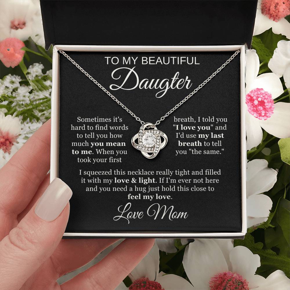 A hand holds an open jewelry box with a To My Beautiful Daughter Cubic Zirconia Love Knot Necklace, featuring a heart-shaped pendant. The black message card has a heartfelt note from mom. Flowers surround the box, perfect for graduation or birthday gifts.