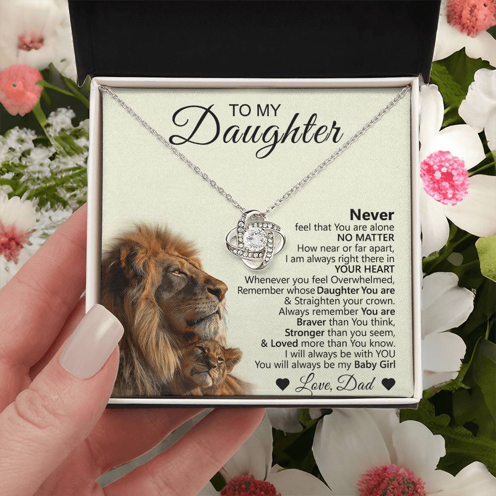 A hand holds the To My Daughter – Love Knot Necklace with Lion & Cub Message Card gift box, featuring a lion and cub illustration, touching father-to-daughter message, and surrounded by pink and white flowers.