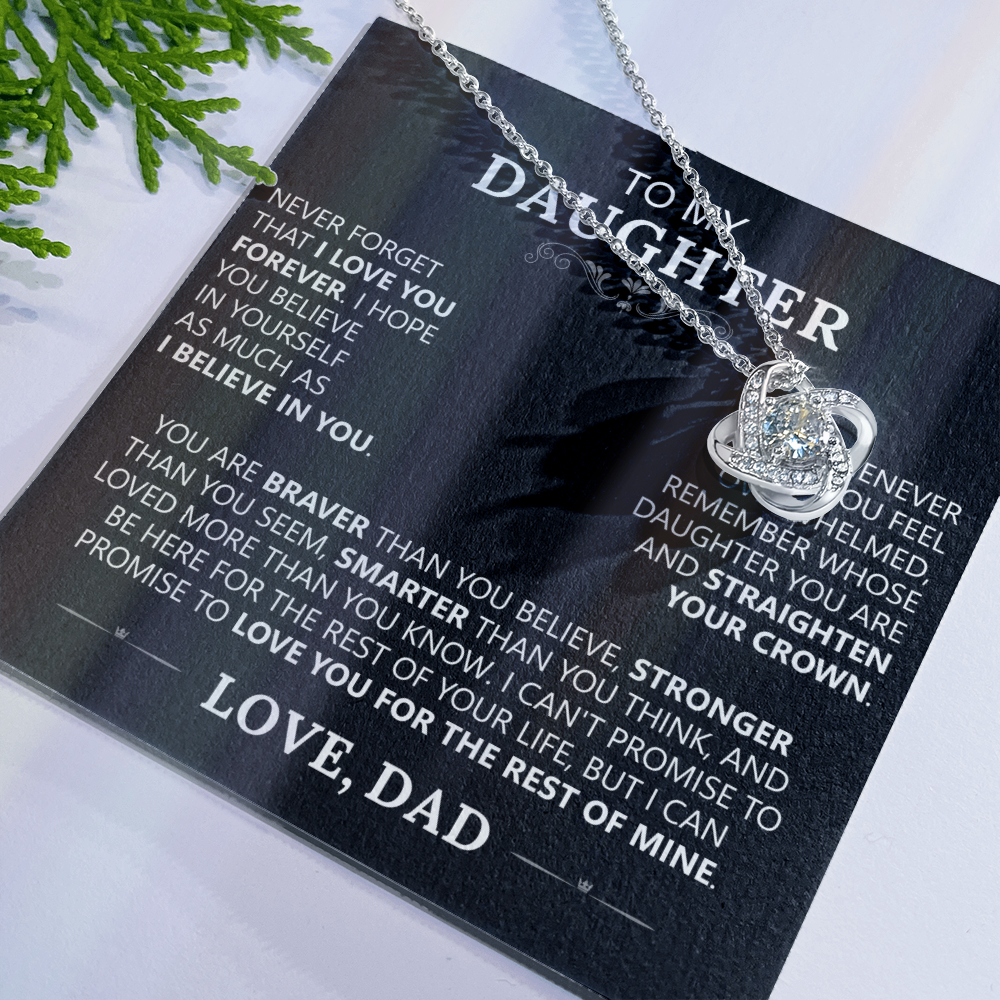 The “To My Daughter - Love Knot Necklace” features a silver heart-shaped pendant on a card with an inspiring father-to-daughter message in white text on a black background, accented by greenery in the corner, symbolizing their unbreakable bond.