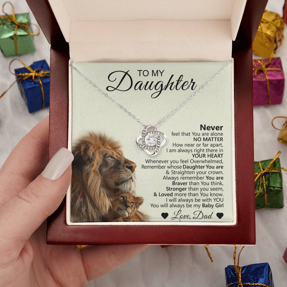 A hand presents the To My Daughter – Love Knot Necklace with Lion & Cub Message Card in an open jewelry box. The lid displays a heartfelt note from Dad, featuring a lion and cub image, with colorful gift boxes in the background.