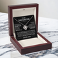 The To My Daughter - Love Knot Necklace comes in a red and cream box, displayed on marble. Behind it is a heartfelt card from dad to daughter, celebrating their unbreakable bond with words of love and support.
