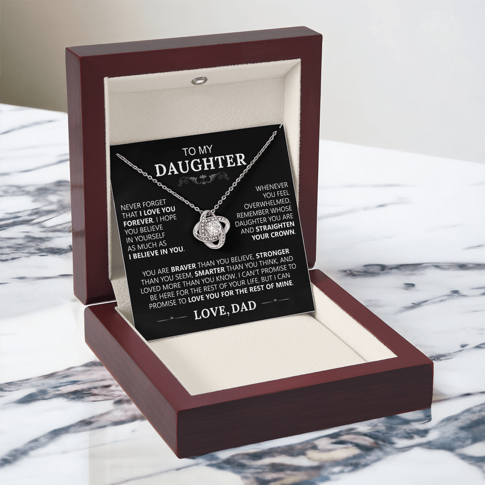 The To My Daughter - Love Knot Necklace comes in a red and cream box, displayed on marble. Behind it is a heartfelt card from dad to daughter, celebrating their unbreakable bond with words of love and support.