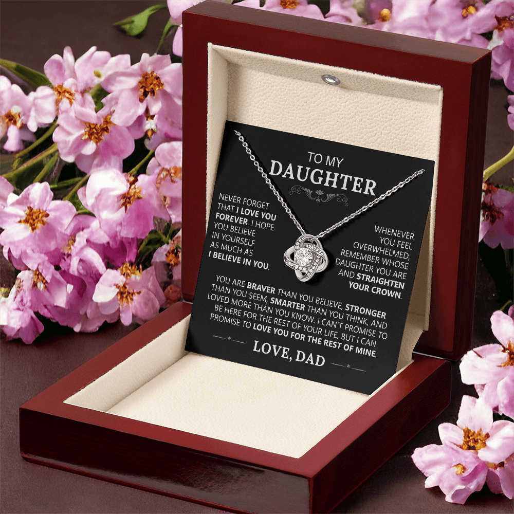 The To My Daughter - Love Knot Necklace comes in a wooden jewelry box with a heartfelt message from a father on a card and pink flowers around it—an ideal father-daughter gift displayed on a dark surface.