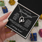 A hand holds an open box revealing the To My Beautiful Granddaughter - Forever Love Necklace, featuring a heart-shaped pendant with cubic zirconia. Against a black backdrop with a heartfelt message card, colorful gift boxes are blurred in the background.