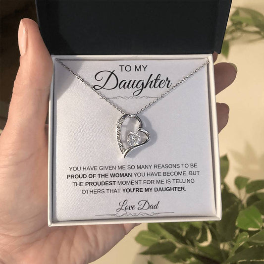 A hand holds an open box with the To My Daughter: Forever Connected Message Card & Love Knot Necklace Set inside. The message card conveys a fathers pride and love for his daughter. Green plant leaves appear in the background.
