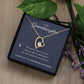 The To My Granddaughter - Forever Love Necklace with Heartfelt Message Card features a heart-shaped pendant with CZ crystal in a black box. Its white gold finish sparkles alongside a loving message, surrounded by stars and a crescent moon, against green leaves and purple flowers.